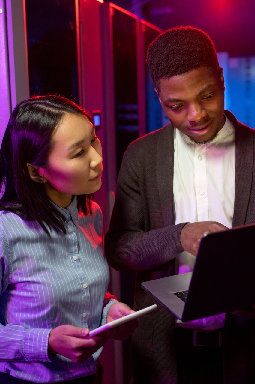 Busy young multi-ethnic colleagues using laptop while monitoring network performance in neon server room