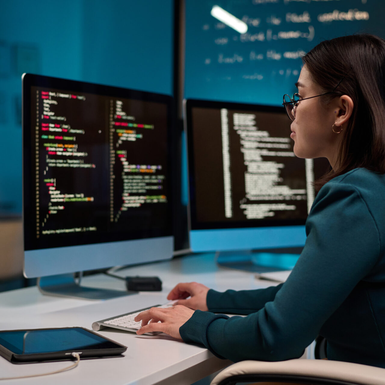 Portrait of young woman as software developer writing code while using computer at office workplace in blue light copy space